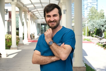 A smiling surgeon with dark hair and a beard stands in a covered walkway, wearing a blue scrub top and a watch. He poses with one hand on his chin and the other crossed. The background shows columns and greenery.